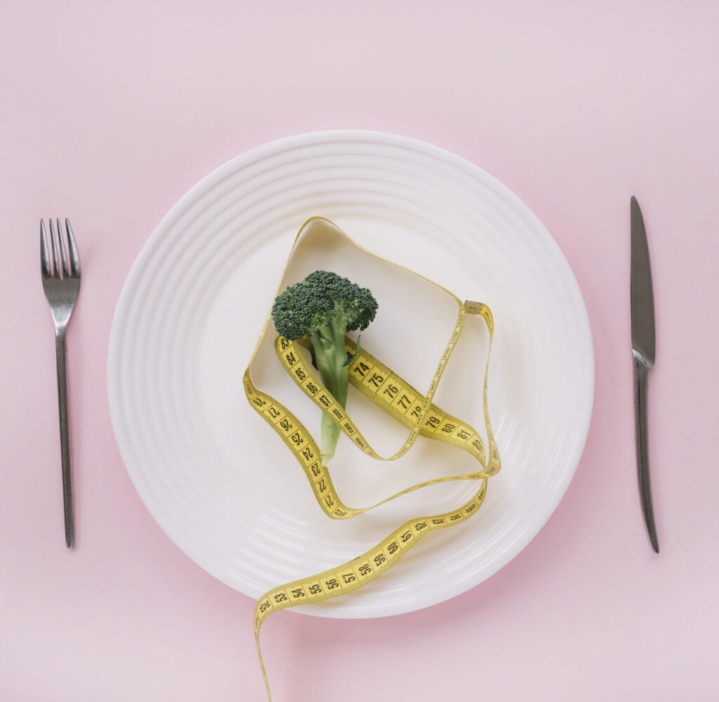 Image of a piece of broccoli on a plate with a measuring tape wrapped around it, which can be used to portion control meals for intermittent fasting.